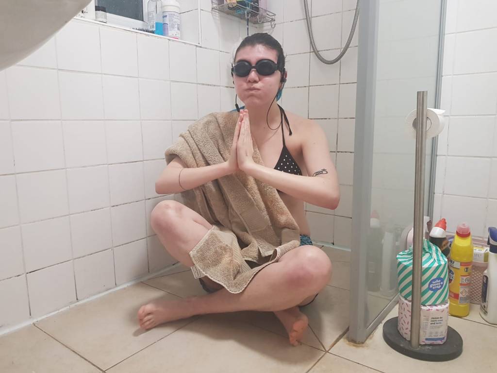 Ellie, a tall white woman, sits cross legged in her white-tiled wet room - clasping her hands together and holding her breath as if she is about to dive. She has a light brown towel draped across her shoulder, and is wearing goggles and a black & white polkadot bikini. 