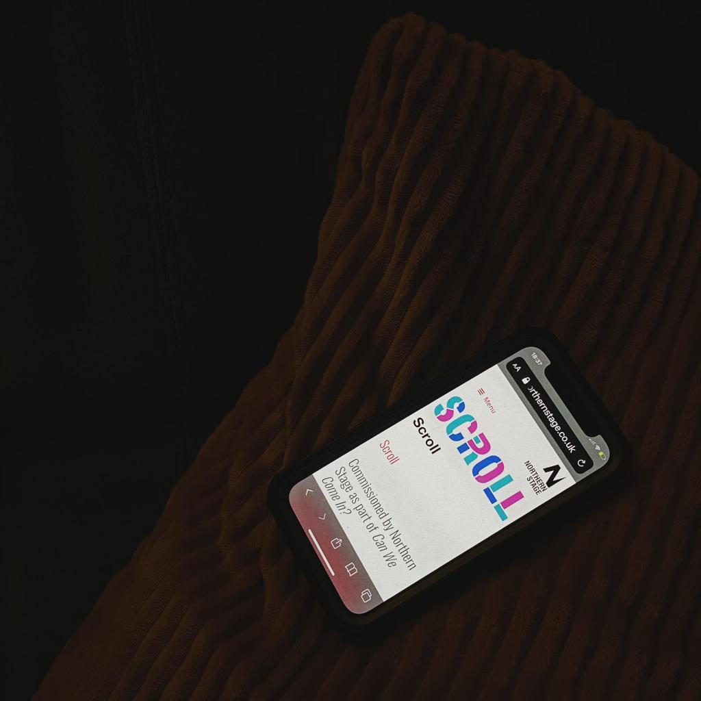 We see a dark room, the only thing visible is the texture of a cushion and the bright screen of a mobile phone. The phone is showing the 'SCROLL' homepage, with the word emblazoned across the screen in multi-coloured, bold text.