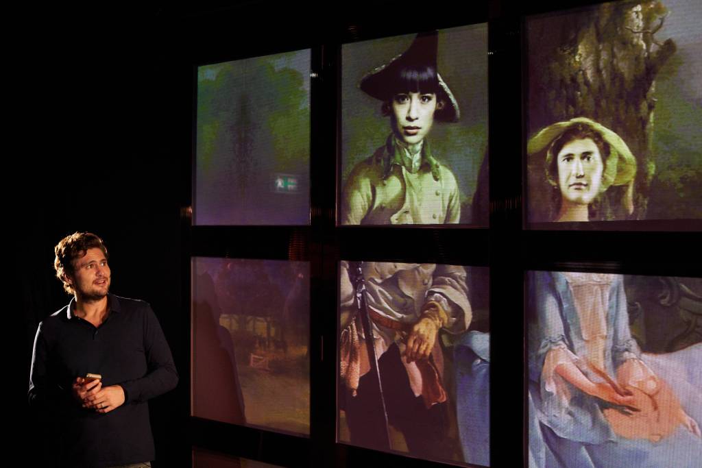 A photo from a live performance of Rich Kids. Javaad stands to the side in a black shirt, looking at a large set of screens arranged in a grid. An image is spread across them, depicting a historical painting. The figures faces have been swapped for the faces of Javaas and Peyvand, like an instagram 'face swap' filter.
