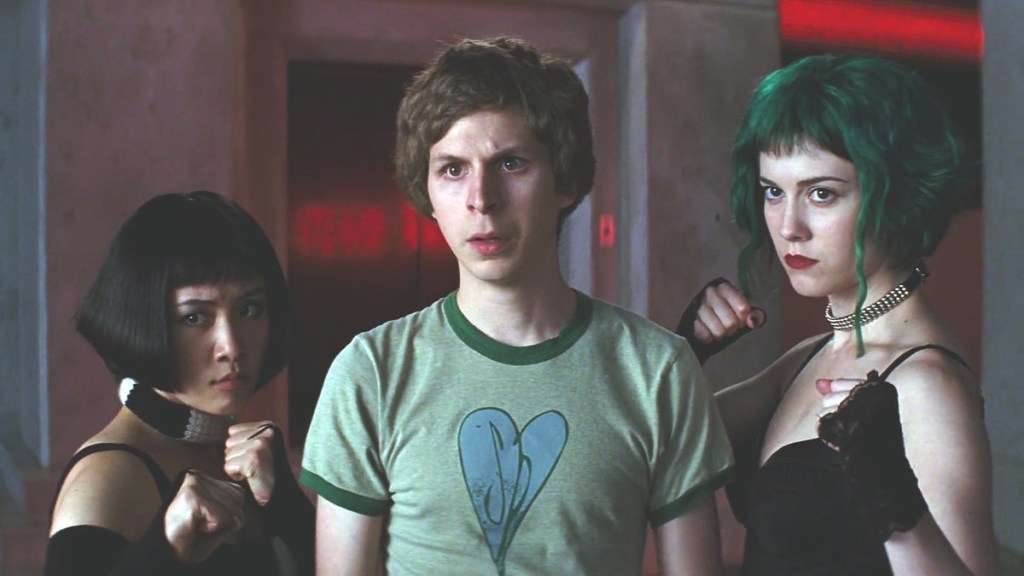 Scott Pilgrim (a thin white man with tousled mousey hair) is center to the image, eyebrows furrowed. On his right hand side is Knives, a short Asian woman with a harsh black bob, and on his left is Ramona, a tall white woman with choppy green bob. Both Knives and Ramona have their fists raised in a fighter-stance, ready for a tussle.