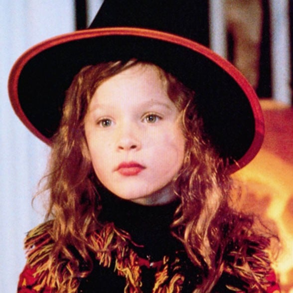 Dani is dressed as a witch for Halloween. She has long wavy brown hair, green sparkling eyes and a black hat with a red trim on her head.