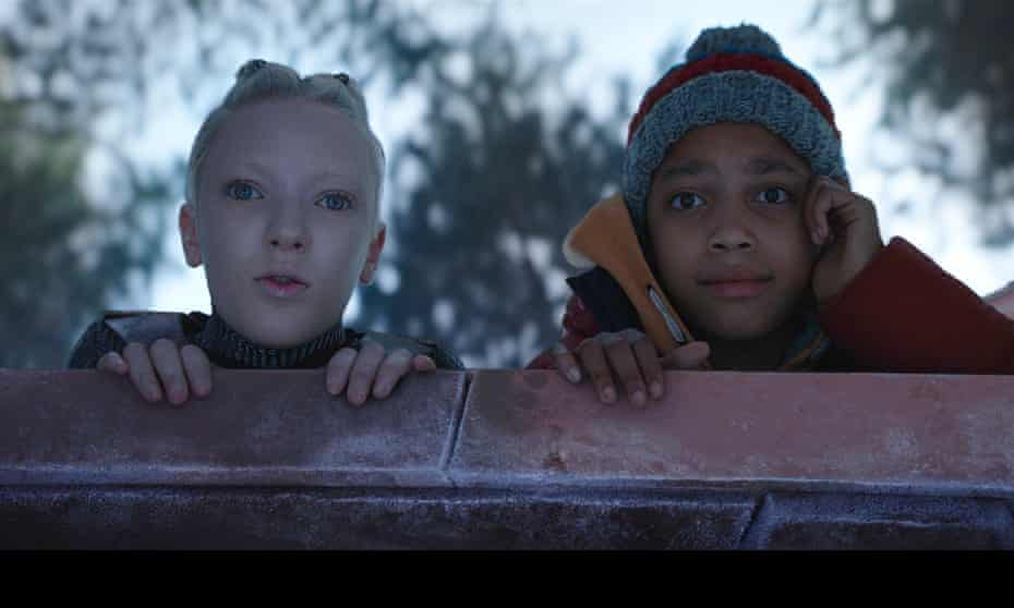 Two children peer over a ledge, looking in to the distance. One is a black child in a woolly hat, the other is a pale, white child. She is an alien. Behind them are trees. 