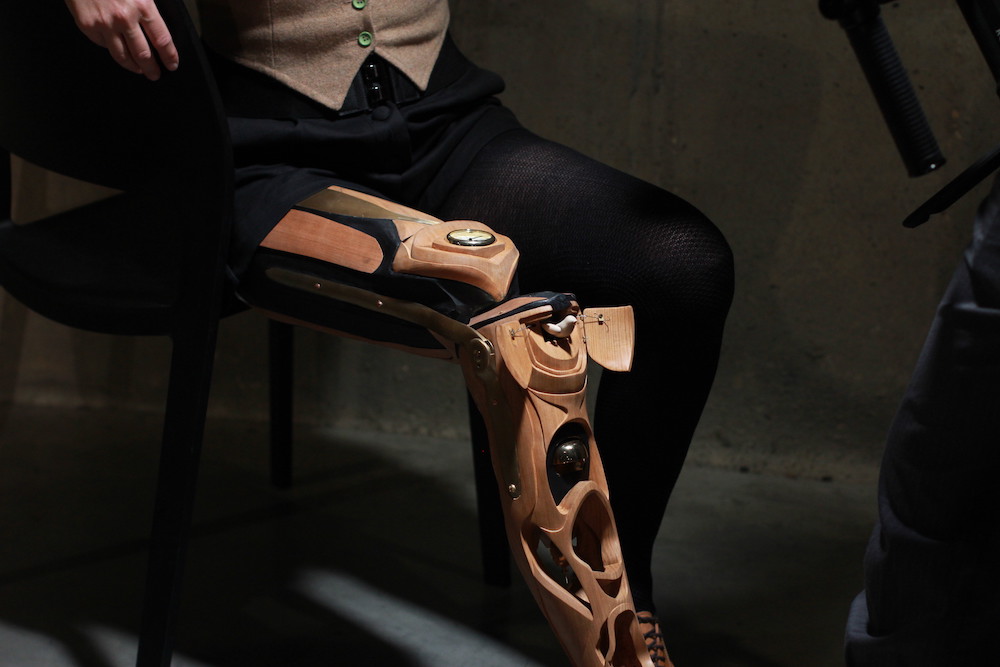 A wooden prosthetic leg with intricate carving is lit up by a spotlight, with the rest of the image in darkness. It has a watch face set into it, and a small wooden cuckoo clock bird coming out of a door. 