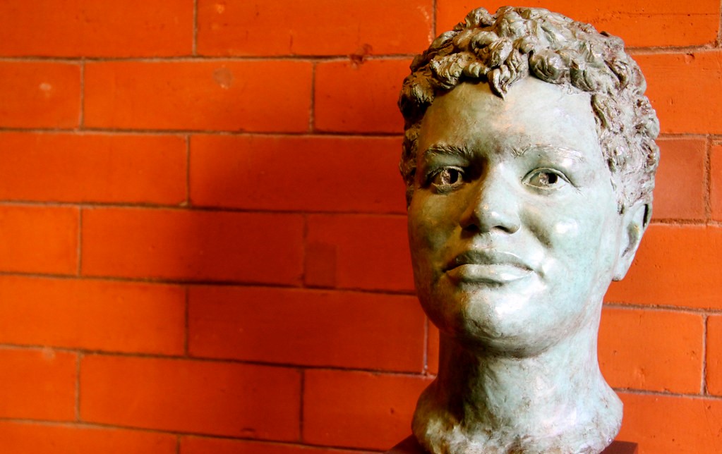 A grey stone statue of Jackie Kay's head and neck is in the foreground, with a very good likeness. It is in front of a bright orange brick wall. 