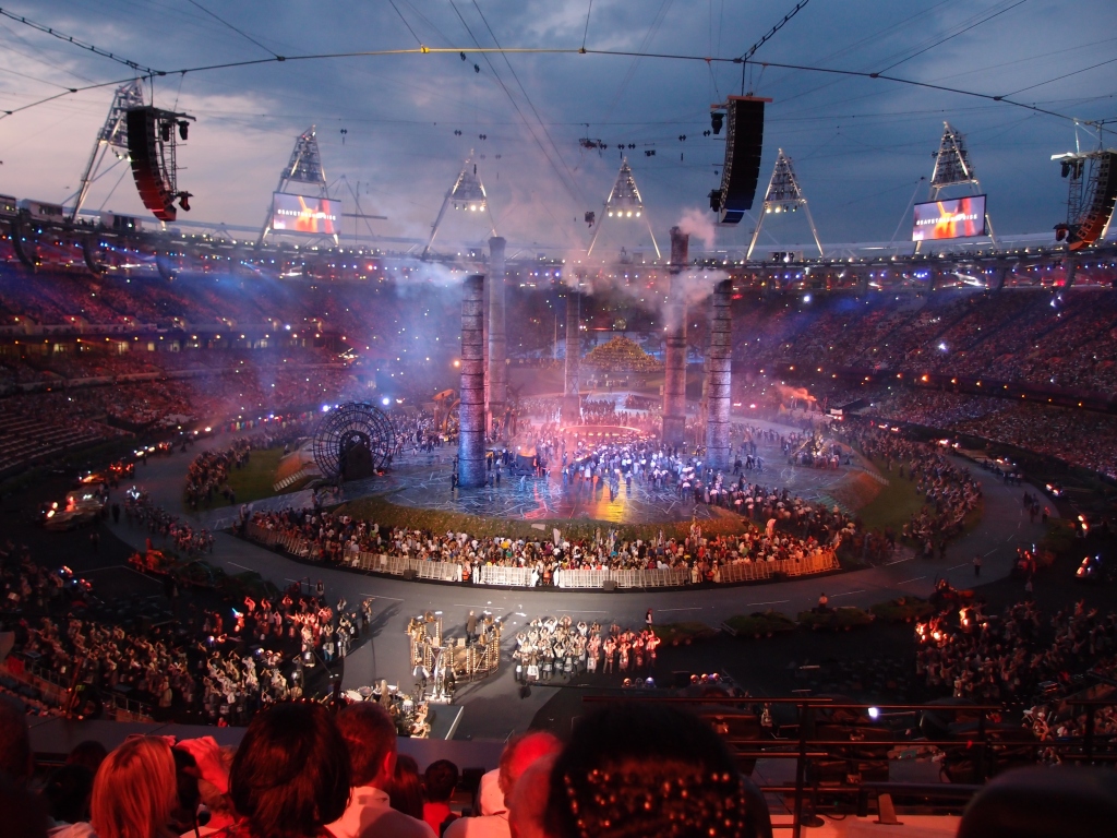 A distant picture of the packed Olympic stadium, with hoardes of performers gathered around smoking chimneys. Large close-up screens are suspended above the stadium on wires, as well as speaker stacks. The stadium is lit in blue, purple and pink (which feels unintentionally bisexual). 